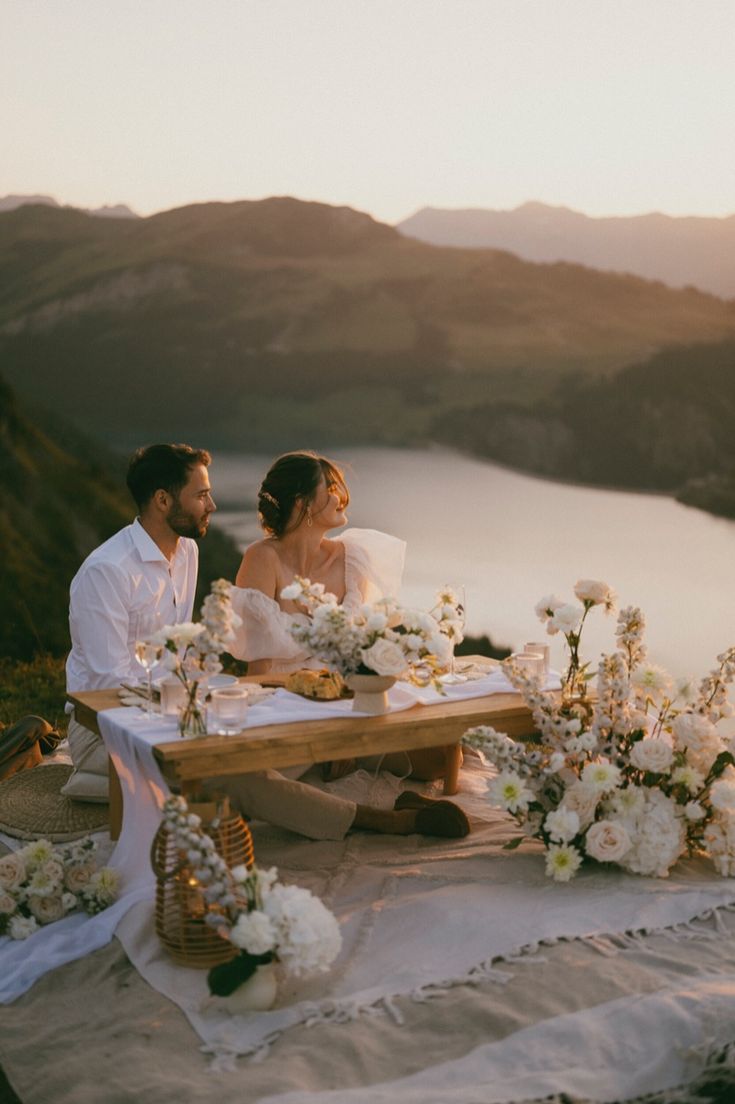Couple seated at a flower-filled lakeside picnic table at golden hour with mountain peaks behind